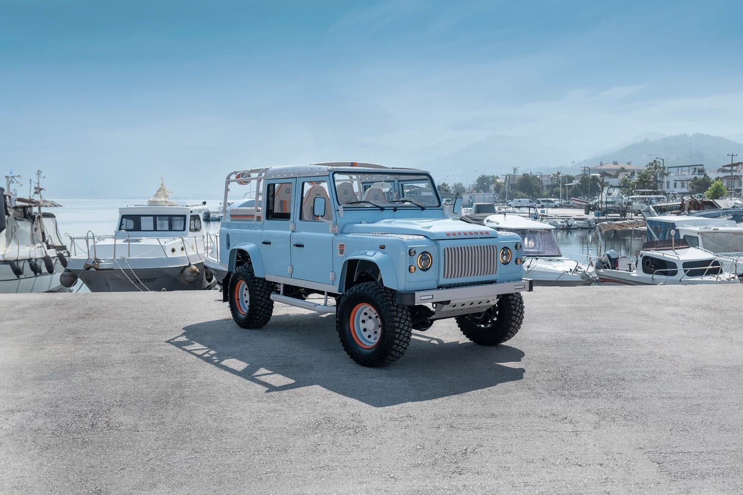 A restored classic Defender 110 in Gulf Runner Blue, photographed in profile against a private yacht backdrop, emphasizing the iconic boxy silhouette